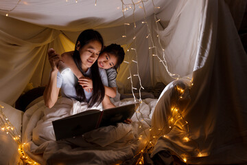 Obraz na płótnie Canvas Family concept. Mother and child daughter reading book with flashlight together in children tent before bedtime. Happy mother read story book to her daughter in bed sheet tent.