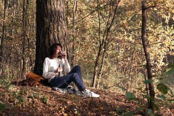 Side view of a traveler young women drinks tea from thermos bottle sitting in the forest in autumn near a tree. Girl drinking tea during hike in forest in autumn