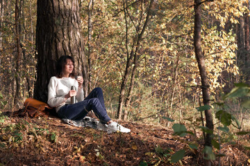 Fototapeta premium Side view of a traveler young women enjoying a hot drink during hiking sitting on the ground near the tree in park. Girl drinking tea during hike in forest in autumn