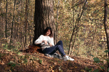 Fototapeta na wymiar Side view of a traveler young women pouring tea from thermos bottle sitting in forest in autumn. Concept of safe travel. Girl drinking tea during hike in forest in autumn