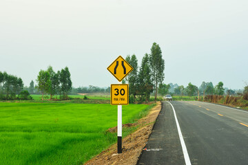 Traffic signs warn of speed limits because of the narrow road ahead on a paved road that cuts through the middle of green fields