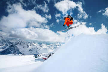 Naklejka na ściany i meble Man skier athlete makes a jump in flight on a snowy slope against the backdrop of a blue sky of mountains and clouds. Freeride and extreme skiing for men