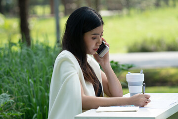 Asian business woman with long hair In a gray-blue suit Sitting on the table in the park Working and talking on the mobile phone Smile happily