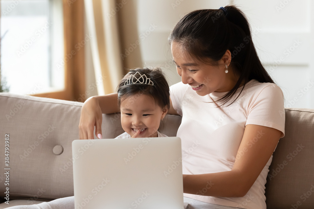 Wall mural happy asian young mother and little daughter using laptop together, looking at screen, sitting on co