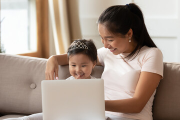Happy Asian young mother and little daughter using laptop together, looking at screen, sitting on cozy couch at home, smiling mum and cute toddler girl child wearing princess diadem having fun