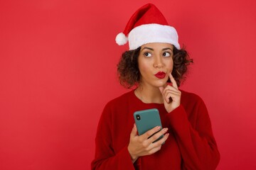 Image of a thinking dreaming young beautiful woman wearing Christmas hat posing isolated over bright wall background using mobile phone.