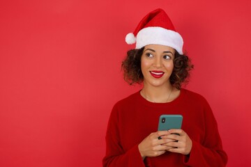 Image of a happy young beautiful woman wearing Christmas hat posing isolated over red background listening to music with earphones using mobile phone.