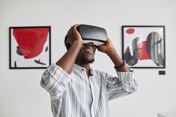 Graphic waist up portrait of smiling African-American man wearing VR gear while enjoying immersive experience at modern art gallery exhibition, copy space