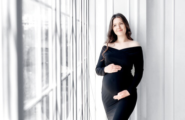 beautiful young pregnant woman in dark black dress stands near the window