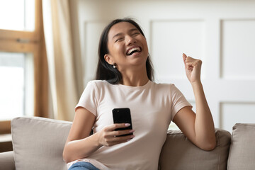 Overjoyed young Asian woman holding phone, celebrating success, showing yes gesture, sitting on...