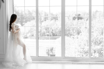 beautiful young pregnant woman in white dress stands near the window black and white photo