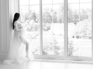 beautiful young pregnant woman in white dress stands near the window