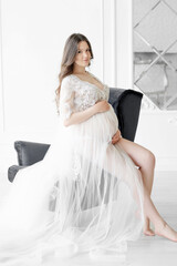 beautiful young pregnant woman in long white dress and sitting on an armchair