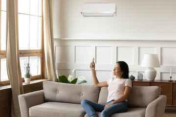 Smiling Asian young woman using air conditioner cooler system remote controller, sitting on cozy...