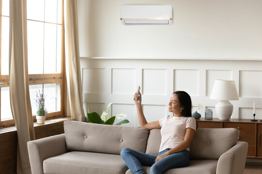 Smiling Asian Young Woman Using Air Conditioner Cooler System Remote Controller, Sitting On Cozy Sofa At Home, Relaxed Girl Switching, Setting Comfort Temperature In Living Room, Enjoy Fresh Air