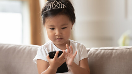 Close up cute little girl wearing princess diadem using smartphone, pretty toddler child looking at screen, playing mobile device game, watching cartoons online, sitting on cozy couch at home © fizkes