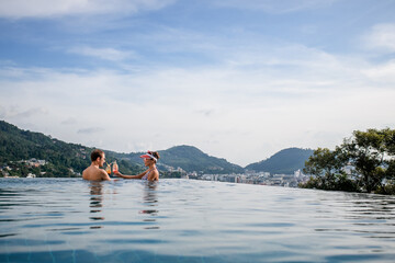 Luxurious couple clink glasses with cocktails while in the infinity pool with a charming view.