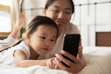 Close up caring Asian mother and little girl using smartphone together, lying on cozy bed in bedroom, curious toddler girl kid touching phone, family enjoying leisure time with gadget at home