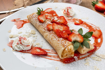 Fresh ripe red juicy strawberries in a golden fried crepe or French pancake drizzled with cream and garnished with mint over a white background