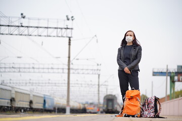 Woman in protective mask waiting for train on platform. Full length of female passenger in protective mask with backpacks standing on railroad platform and waiting for train during coronavirus 