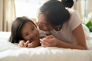 Close up Asian mother kissing smiling adorable little daughter, lying on bed together, family enjoying tender moment, caring loving mum and toddler girl kid enjoying leisure time at home