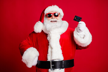 Close-up portrait of his he nice handsome cheerful cheery bearded Santa holding in hand using plastic card store shop cash back cashback isolated bright vivid shine vibrant red color background