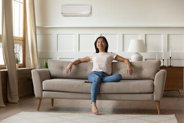 Tranquil calm young Asian woman relaxing in room with air conditioner on wall, sitting on cozy...