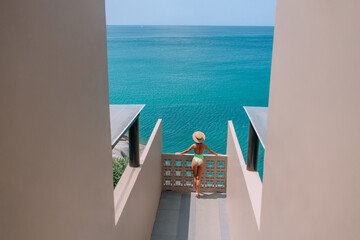 A slender woman in a hat admires the sea horizon while standing on the terrace of her hotel. Side view