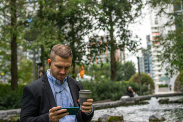 Fototapeta premium Photo of a young and attractive businessman watching a video on his phone with headphones while having a coffee during a work break in the middle of the park. Wearing smart clothes. Entrepreneur.