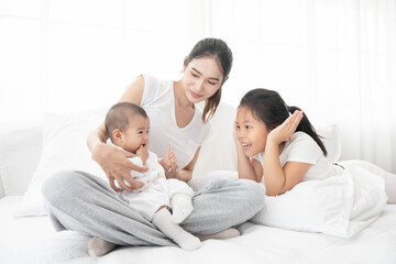 Asian mother and children girl baby laugh together at home. Asia mom holdind infant and play with sister sitting on white bed in living room at house family having fun together.