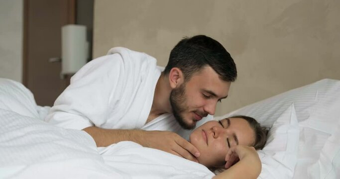 Careful young man kisses girlfriend to wake up in large comfortable bed in modern hotel room in morning at honey moon