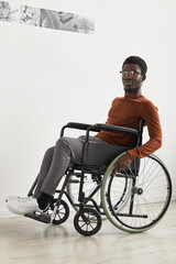 Vertical full length portrait of young African-American man using wheelchair and looking at camera while exploring modern art gallery exhibition