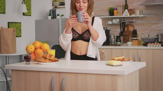 Beautiful young woman in black lingerie drinking coffee smiling looking at camera. Seductive blode lady with tattoos wearing sexy underwear relaxing during morning breakfast sitting in kitchen.