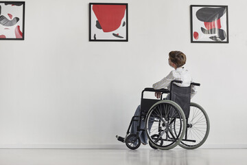 Wide angle portrait of young woman using wheelchair looking at paintings in modern art gallery,...