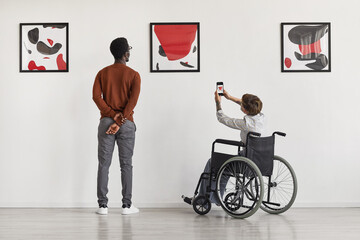Back view portrait of young woman using wheelchair taking photo of artwork while visiting...