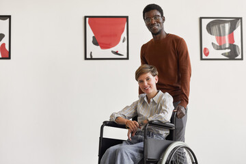 Portrait of young woman wearing mask and looking at camera while visiting modern art museum with African-American man helping her, copy space