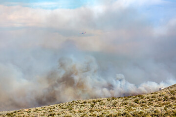 Large orange and white fire tanker flying through heavy clouds of smoke over a wildfire with low visibility