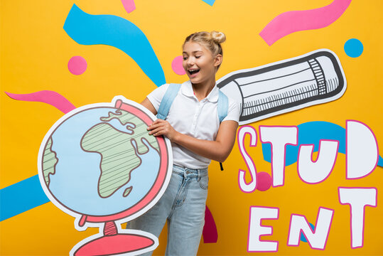 joyful schoolgirl holding globe maquette near student lettering, paper pencil and abstract elements on yellow