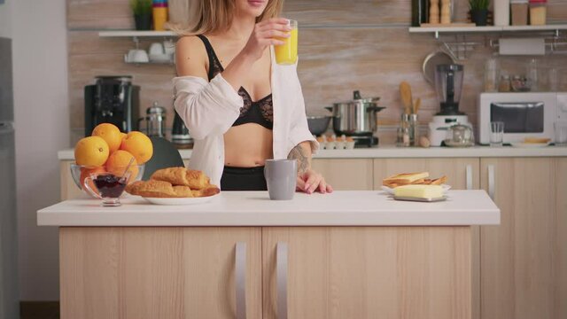Sexy lady drinking orange juice in the morning during breakfast wearing black lingerie. Young seductive blode woman in sexy underwear with tattoos sitting in modern kitchen.