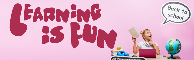 Panoramic shot of kid holding book near globe, gadgets, paper artwork and learning is fun lettering on pink background