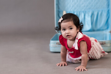 Baby girl sitting with blue suitcase on gray background