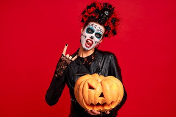 Halloween Party girl. Happy girl with sugar skull makeup, with a wreath of flowers on her head and skull, wearth lace gloves and leather jacket, Holds a big Jack-o-lantern pumpkin isolated on red