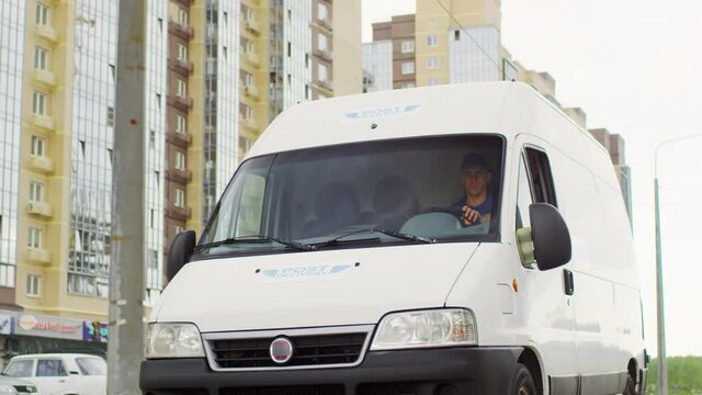 Tracking shot of male courier in blue uniform driving mail delivery van along road in city