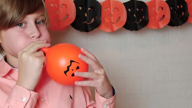 A blond boy smiling Inflating the orange pumpkins balloon with the face of Jack 'O Lantern, celebrate Halloween at home with festive decorations