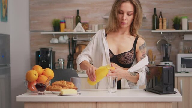 Attractive woman pouring fresh juice in glass for breakfast in kitchen. Young sexy seductive blode lady with tattoos drinking healthy, natural homemade orange juice, refreshing sunday morning