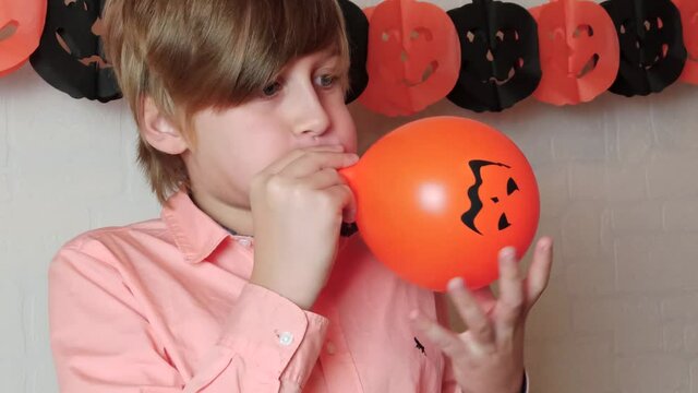 A blond boy smiling Inflating the orange pumpkins balloon with the face of Jack 'O Lantern, get in ready for Halloween at home, celebrate Halloween at home with festive decorations