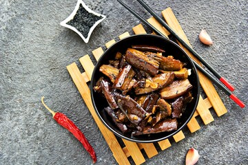 An appetizer or a main dish, fried eggplant in Chinese style with soy sauce and garlic in a black  clay plate on a dark concrete background. Eggplant recipes