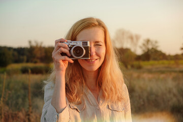 Young blonde caucasian woman is taking pictures on the camera in nature. Camera near the face, woman smiling.