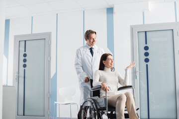 Brunette woman in wheelchair pointing with hand while sitting in wheelchair near doctor in hospital