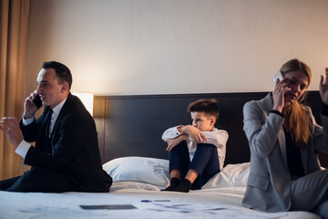 A man and a woman talking on their phones in a hotel room, a boy sitting on the bed between them and listening.